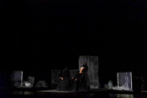 Em um palco escuro, três figuras vestidas de preto estão em cena. Uma mulher de cabelos brancos senta-se, amparada por outra. Ao fundo, painéis de rede cinza compõem o cenário.