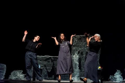 Três atrizes vestidas de escuro em poses sincronizadas de aplausos em cena teatral com fundo preto.