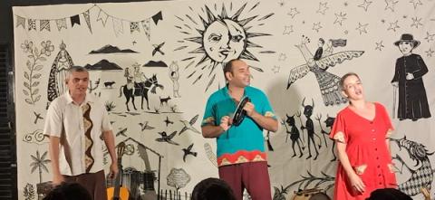 Três artistas com instrumentos em um palco decorado com motivos culturais diante do público.