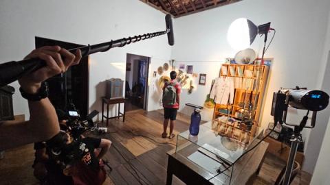 Menino observa instrumentos musicais em parede durante filmagem em sala.