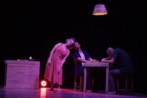 Três pessoas em torno de uma mesa, iluminadas por uma lâmpada pendente em palco escuro.