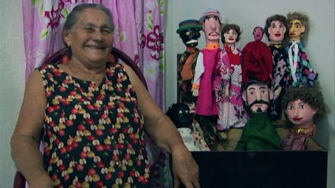 Foto de Dona Dadi sorrindo com bonecos ao lado.