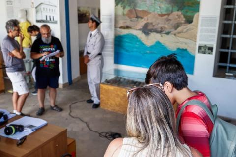 Equipe reunida em salão de museu. Diretor lê roteiro e ator aguarda em pé.
