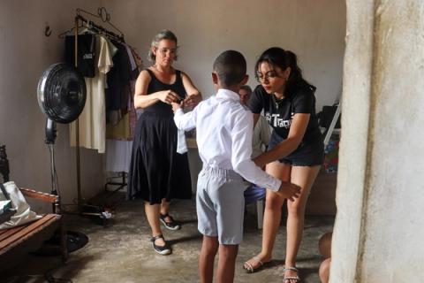 Duas mulheres da equipe de figurino ajustam a roupa de um jovem ator, vestindo-o com uma camisa social branca, em preparação para a gravação.