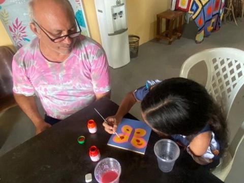 Homem e menina sentados à mesa, pintando com pincéis e tintas coloridas.