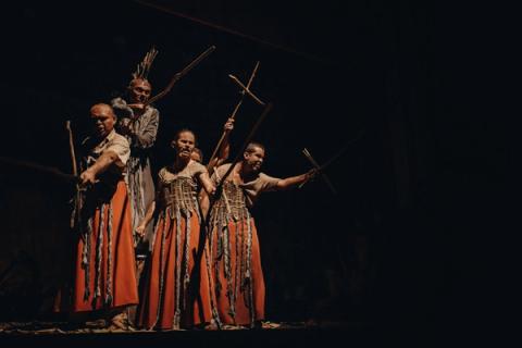 Grupo de atores em cena teatral, vestidos com trajes tradicionais e segurando cruzes de madeira, iluminados contra um fundo escuro.