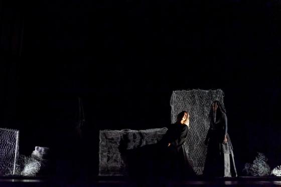 Em um palco escuro, três figuras vestidas de preto estão em cena. Uma mulher de cabelos brancos senta-se, amparada por outra. Ao fundo, painéis de rede cinza compõem o cenário.