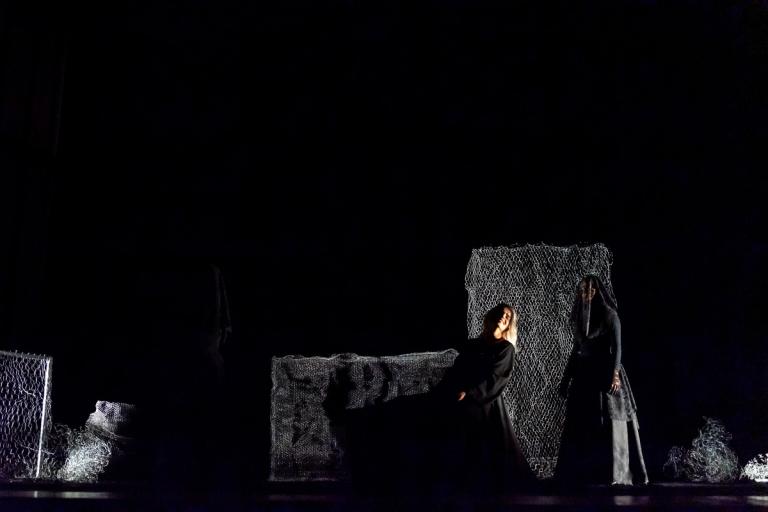 Em um palco escuro, três figuras vestidas de preto estão em cena. Uma mulher de cabelos brancos senta-se, amparada por outra. Ao fundo, painéis de rede cinza compõem o cenário.
