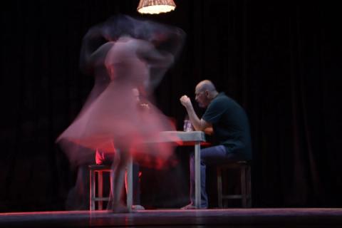 Bailarina em vestido vermelho, em cena com lâmpada pendente e fundo escuro.