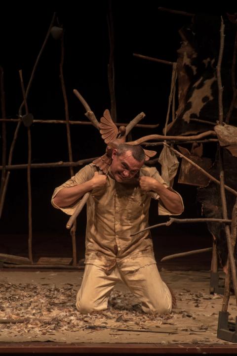 Ator agachado, expressão intensa, com objeto sobre os ombros.