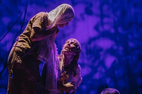 Duas figuras se encaram intensamente em cena teatral, iluminadas por luz roxa.