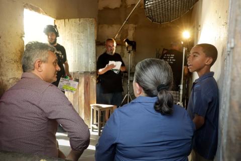 Bastidores de uma cena em um ambiente interno, com o diretor e a equipe ao fundo, enquanto atores, incluindo um menino, estão em posição.