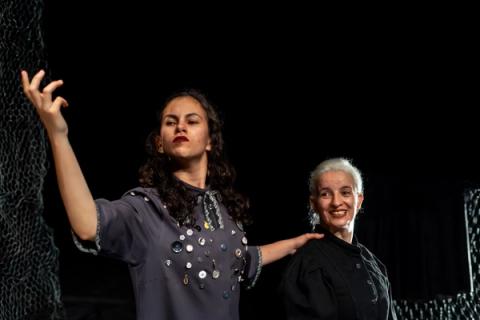 Mulher de vestido cinza com botões gesticula com a mão para o alto, com expressão séria. Atrás dela, uma mulher de cabelos brancos sorri, com a mão em seu ombro.