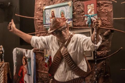 Homem com chapéu e óculos, segurando faca e colar, em cena teatral.