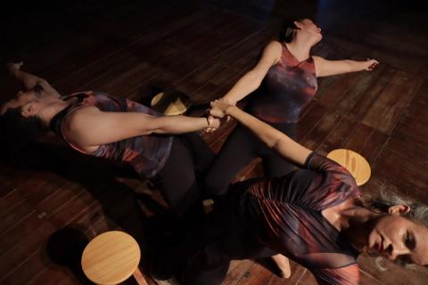 Três bailarinos em movimento, com um segurando a mão de outro, ao lado de banquetas.