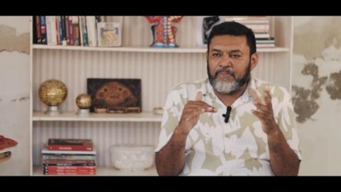 Homem falando, com fundo de estante de livros e objetos decorativos.