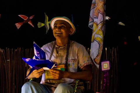 Ator sentado, segurando um pássaro de papel, expressão alegre.