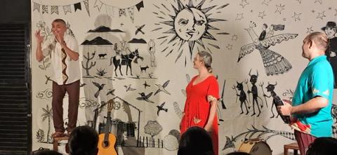 Três artistas com instrumentos em um palco decorado com motivos culturais diante do público.