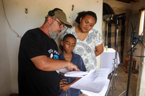 O diretor, usando fones, revisa o roteiro com um jovem ator e uma mulher, enquanto equipamentos de iluminação são vistos ao fundo do set.