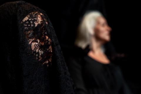 Close-up do rosto de uma pessoa coberto por um véu de renda preta, criando um ar de mistério. Ao fundo, o perfil de uma mulher de cabelos brancos aparece desfocado.