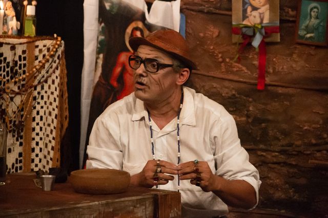 Ator vestindo chapéu e óculos, sentado à mesa em cena teatral.