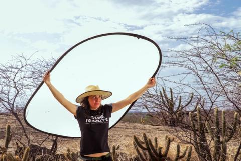 Em uma locação externa com vegetação seca, uma mulher da equipe técnica, de chapéu, segura um grande rebatedor circular para iluminar o set.