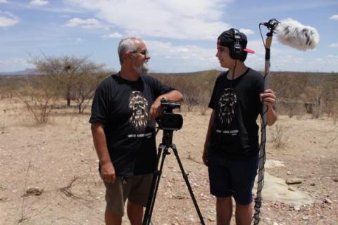 Duas pessoas em um ambiente desértico, uma com microfone e outra com câmera.