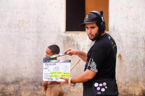 Um homem da equipe de filmagem segura uma claquete do filme "Pemba", preparando-se para iniciar a gravação de uma cena com um menino ao fundo.