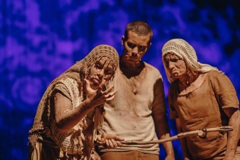 Três atores em cena teatral, com expressões dramáticas e um deles segurando uma vara.