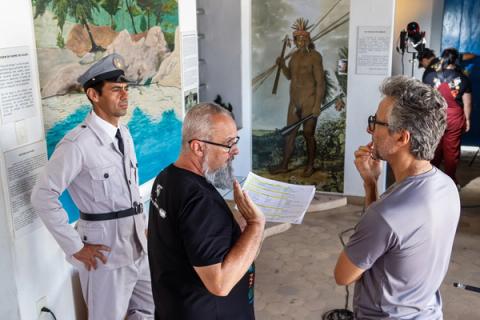 Diretor instrui ator uniformizado em frente a painéis expositivos do museu.