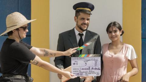 Três pessoas posando com claquete: uma mulher segurando a claquete, um homem em terno e chapéu de capitão, e outra mulher ao lado.