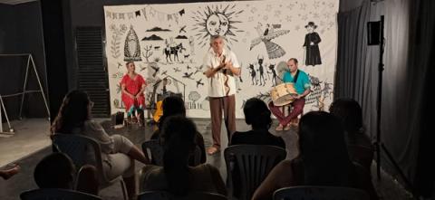 Três artistas com instrumentos em um palco decorado com motivos culturais diante do público.