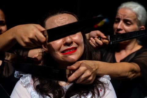 Close de atriz com batom vermelho cercada por pessoas segurando facas próximas ao seu rosto em cena teatral dramática.