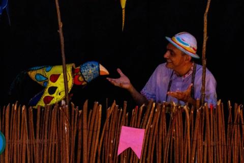 Ator interagindo com marionete de pássaro em frente a cerca de madeira.