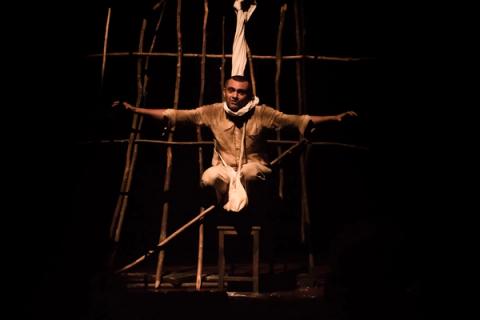 Ator sentado em estrutura de madeira, braços estendidos, expressão intensa, lençol enrolado no pescoço.