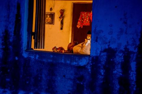 Homem deitado na janela de um ambiente azul, com quadros e cortina colorida ao fundo.