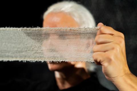 Close-up de uma pessoa de cabelos brancos segurando uma faixa de tecido rústico na frente do rosto, cobrindo parcialmente sua visão e expressão.