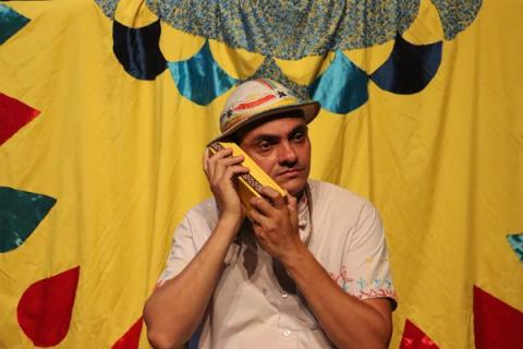 Ator com chapéu colorido, com objeto junto ao rosto em fundo vibrante.