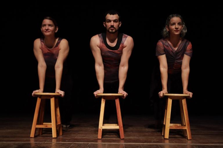 Três bailarinos apoiados em banquetas, com fundo preto e iluminação cênica.