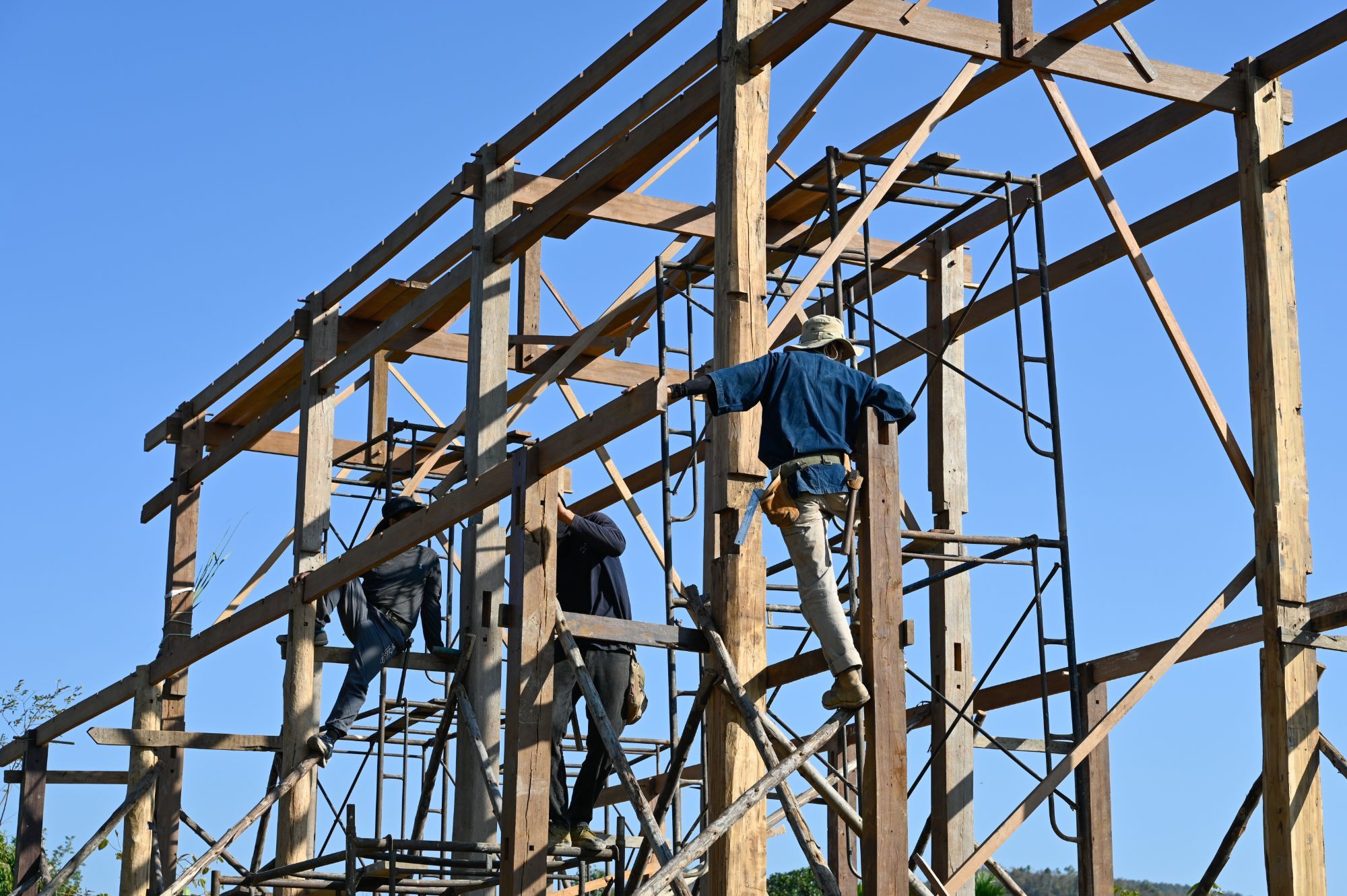 Craftspeople assembling a traditional timber frame on scaffolding under a clear blue sky