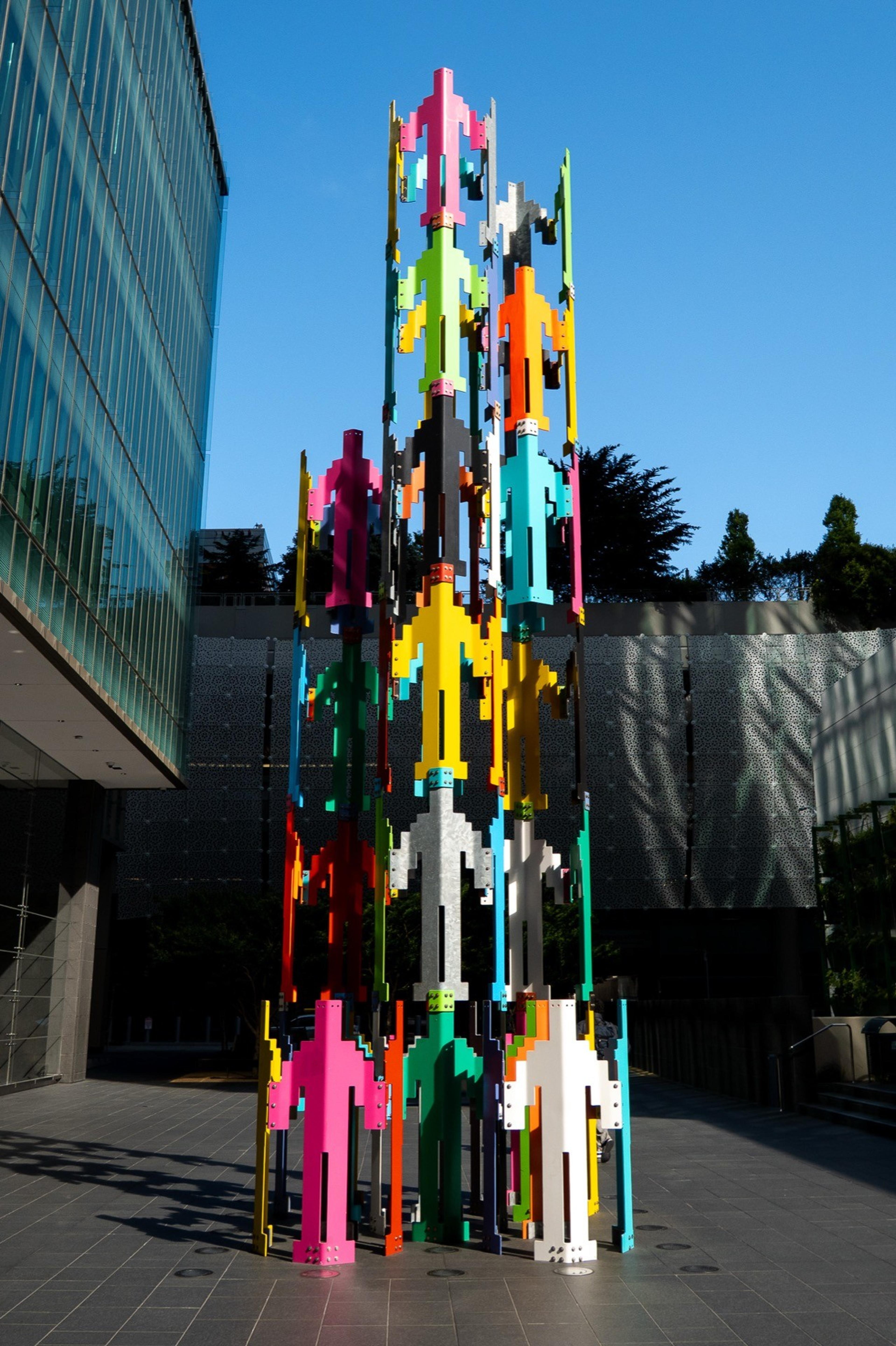 Tall sculpture of stacked, multicolored pixel-style human figures in a plaza under a blue sky