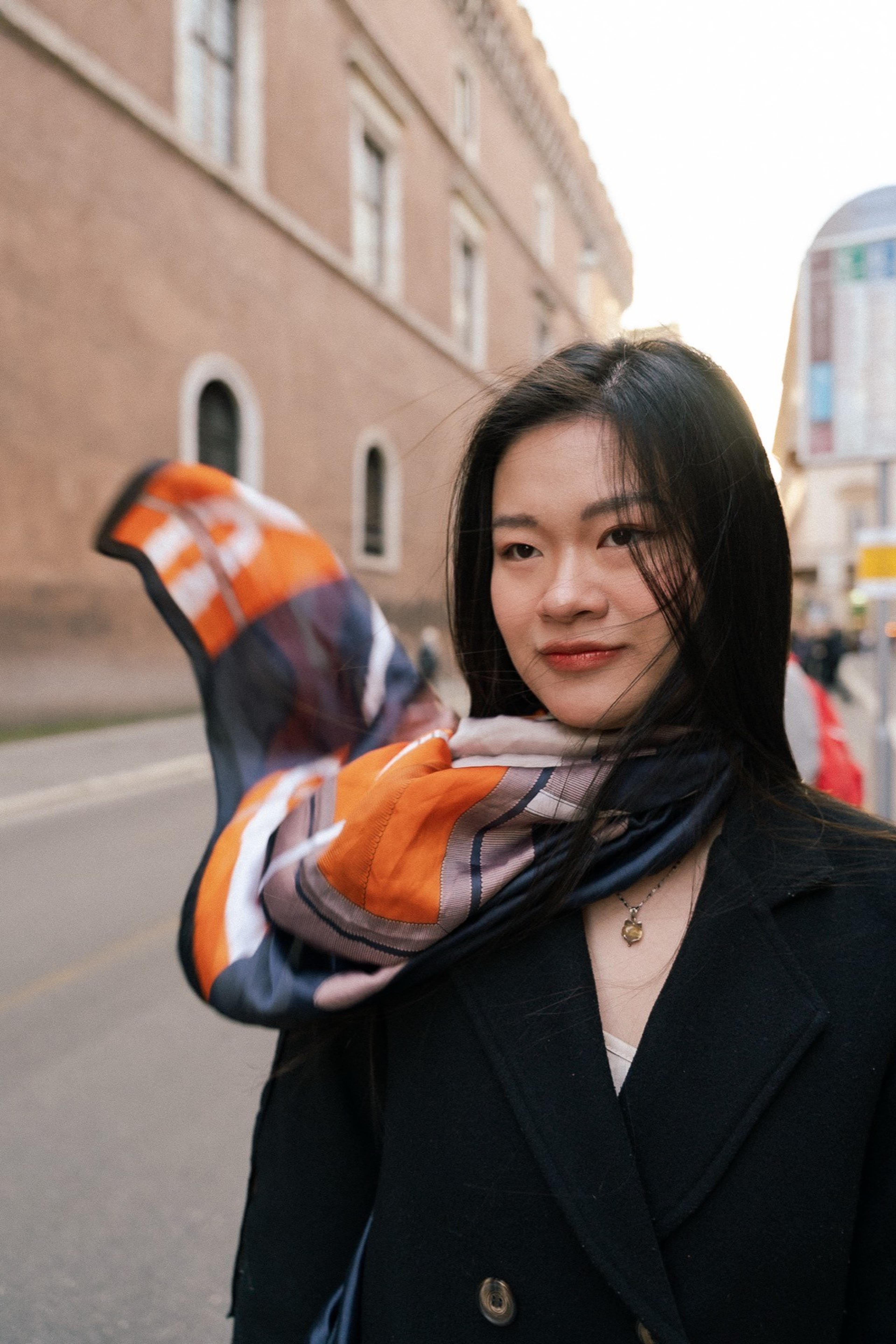Woman in a dark coat with an orange scarf blowing in the wind on a city street