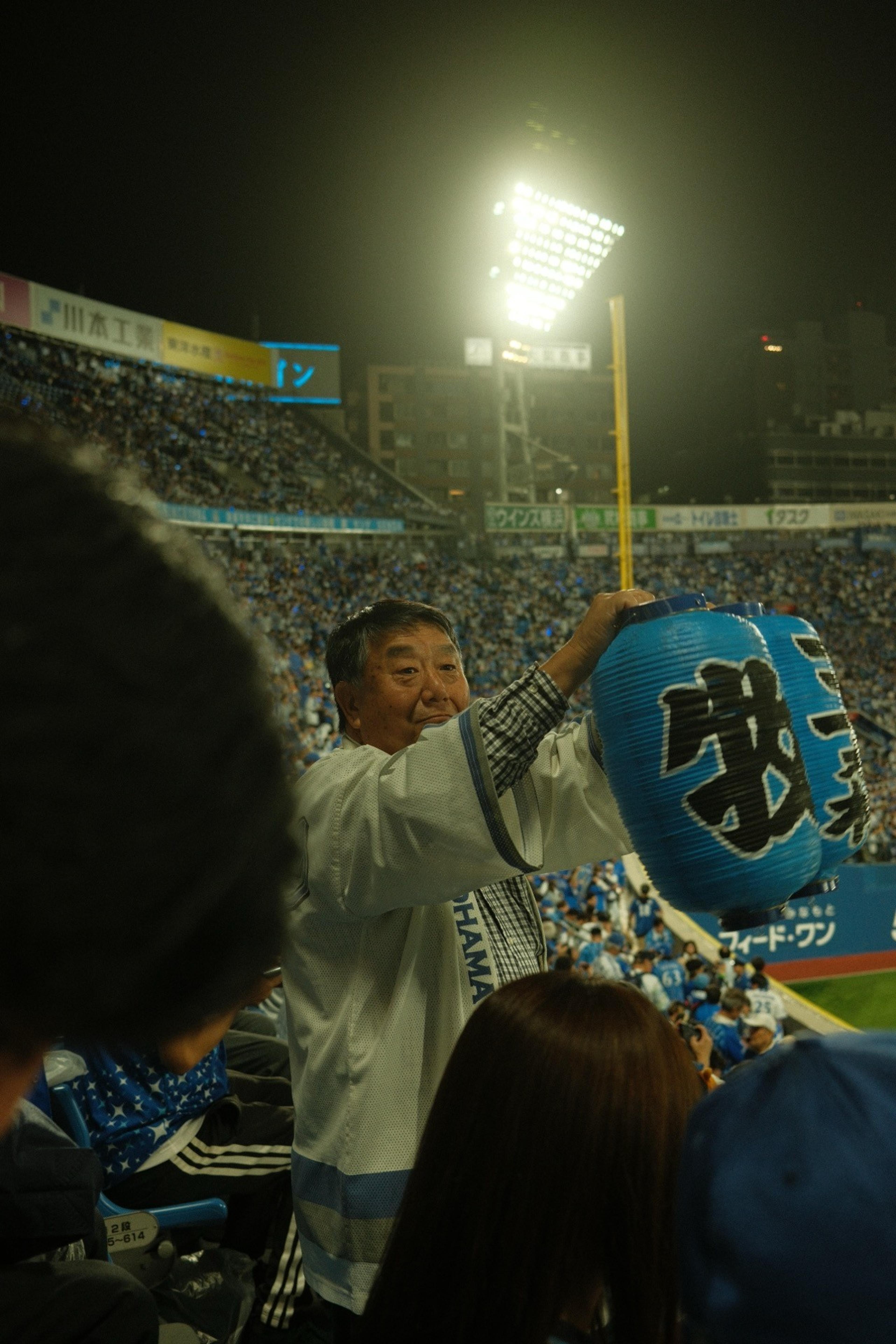 Smiling baseball fan holding up blue paper lanterns in a floodlit stadium at night