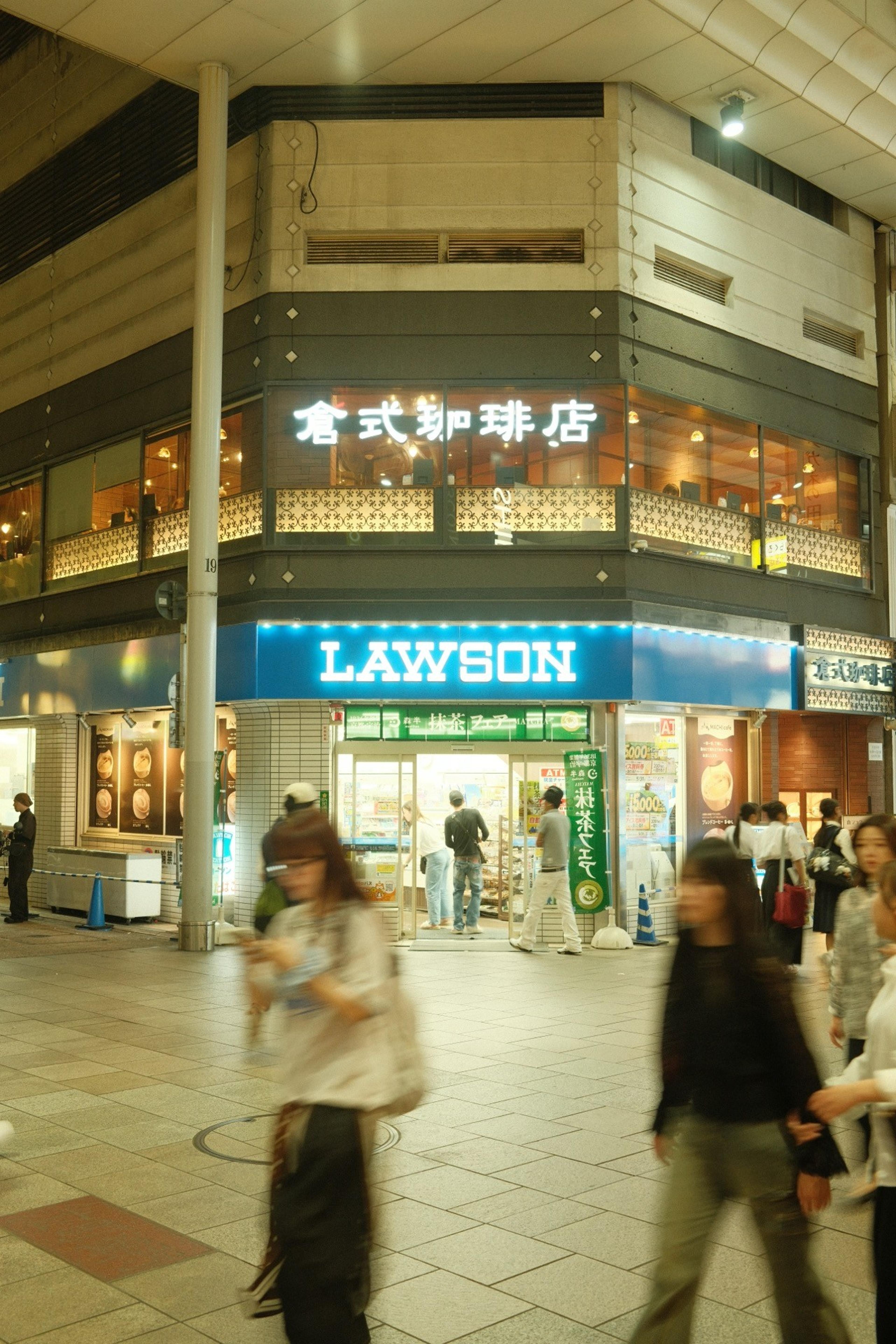 Pedestrians passing a lit Lawson store with a cafe above in an evening shopping arcade