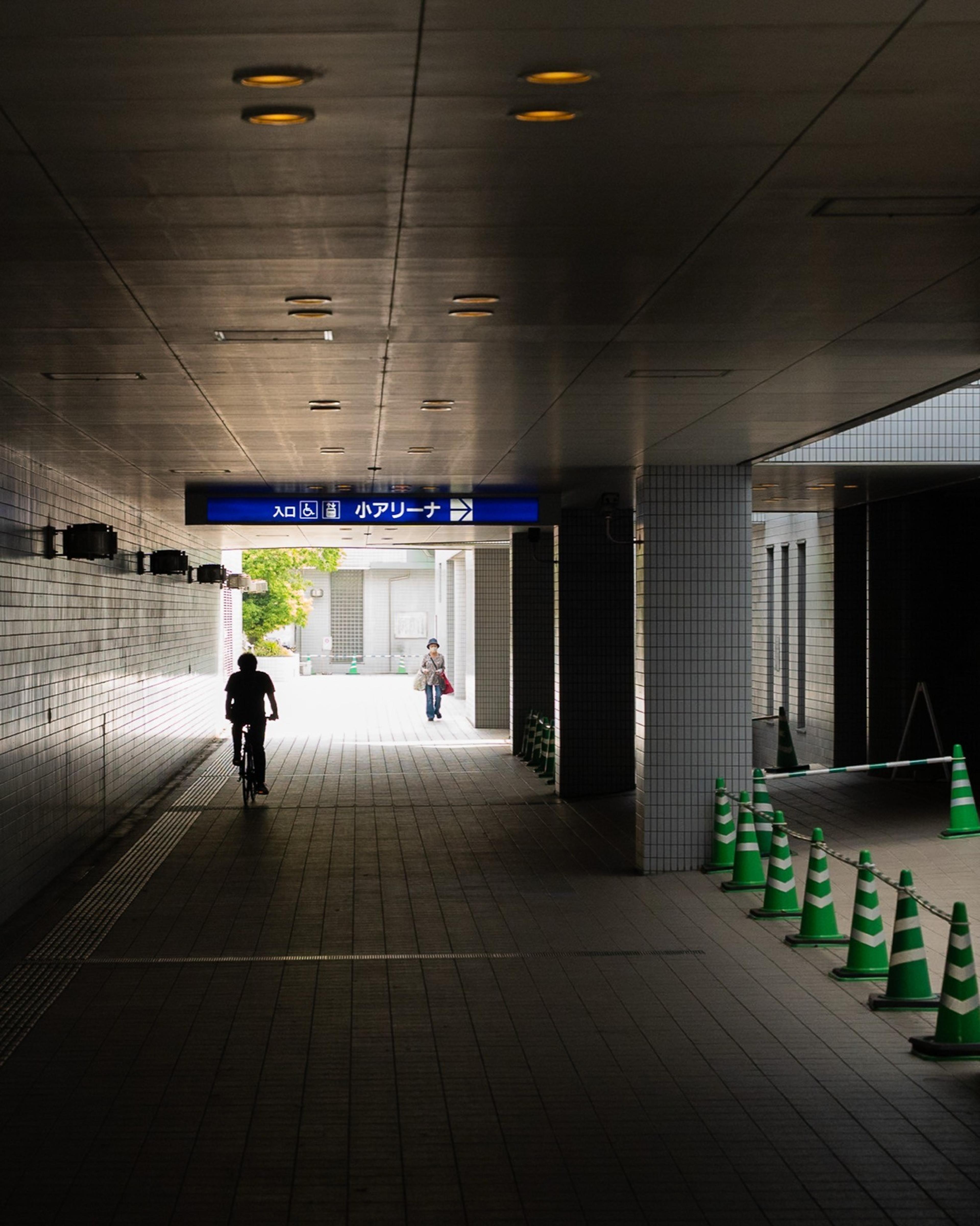 Cyclist in silhouette riding through a tiled underpass toward daylight, green traffic cones aside