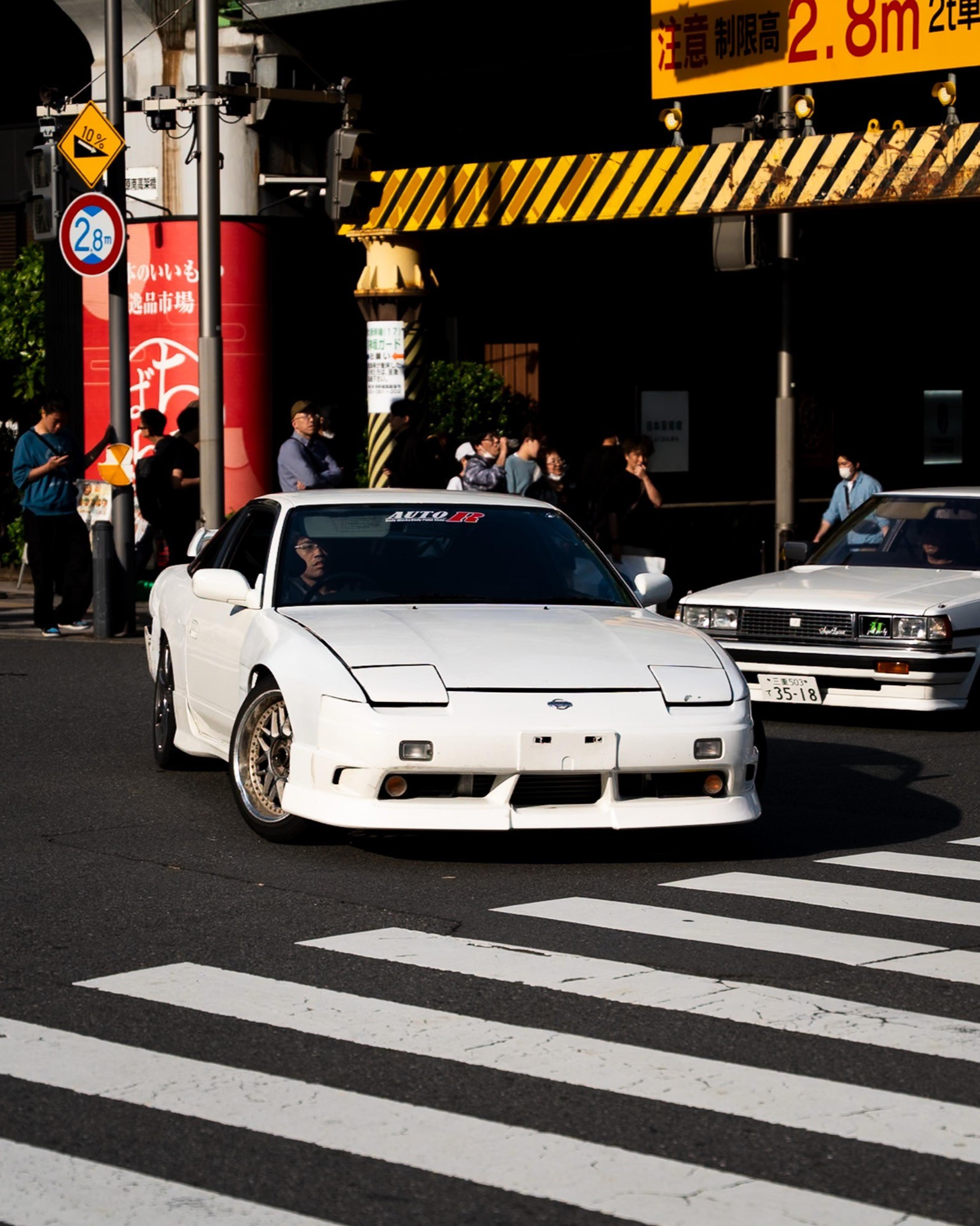 White sports car turning across a crosswalk under a railway bridge as onlookers take photos