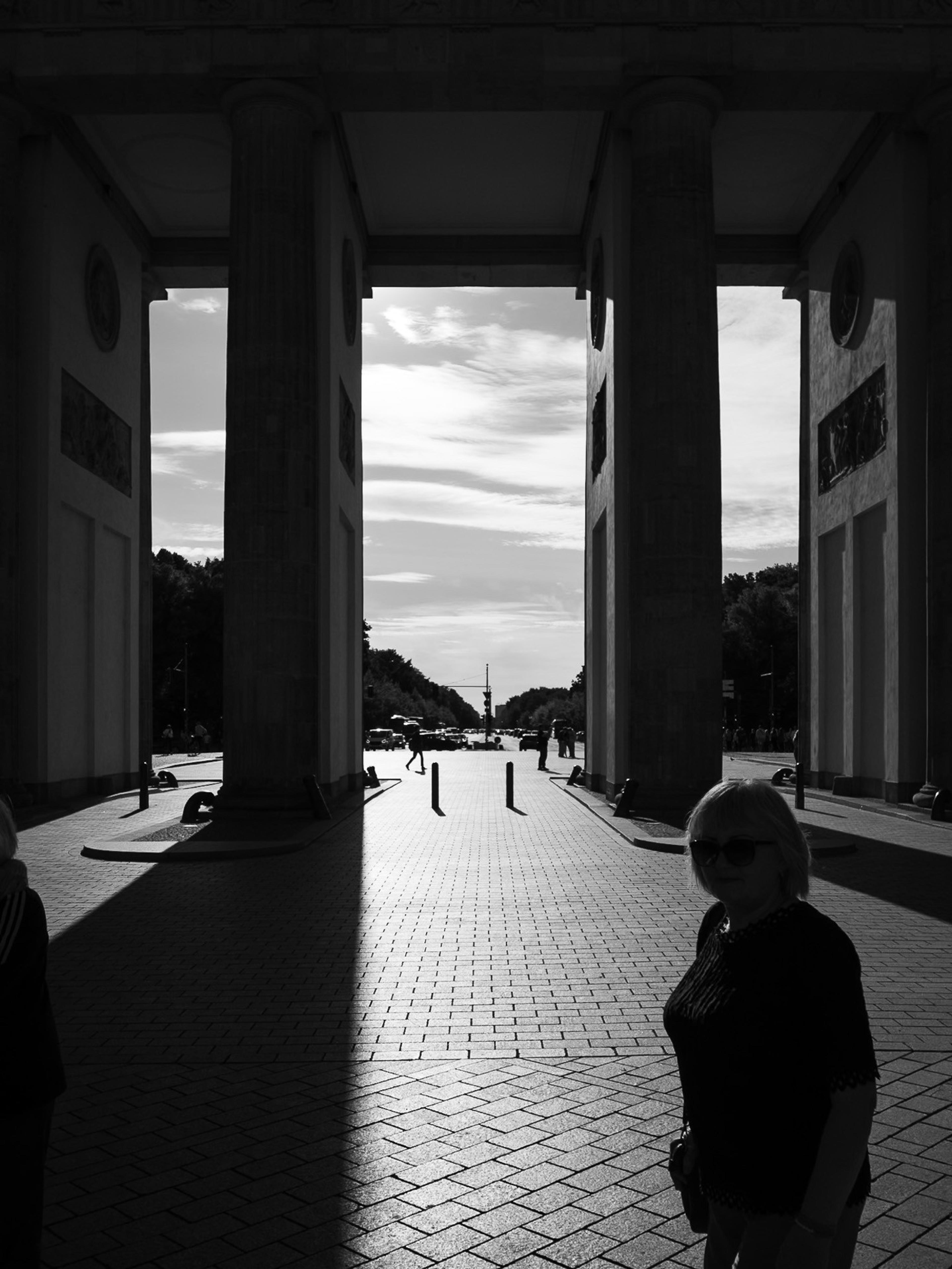 Black and white view through massive stone columns onto a sunlit boulevard, a woman in silhouette