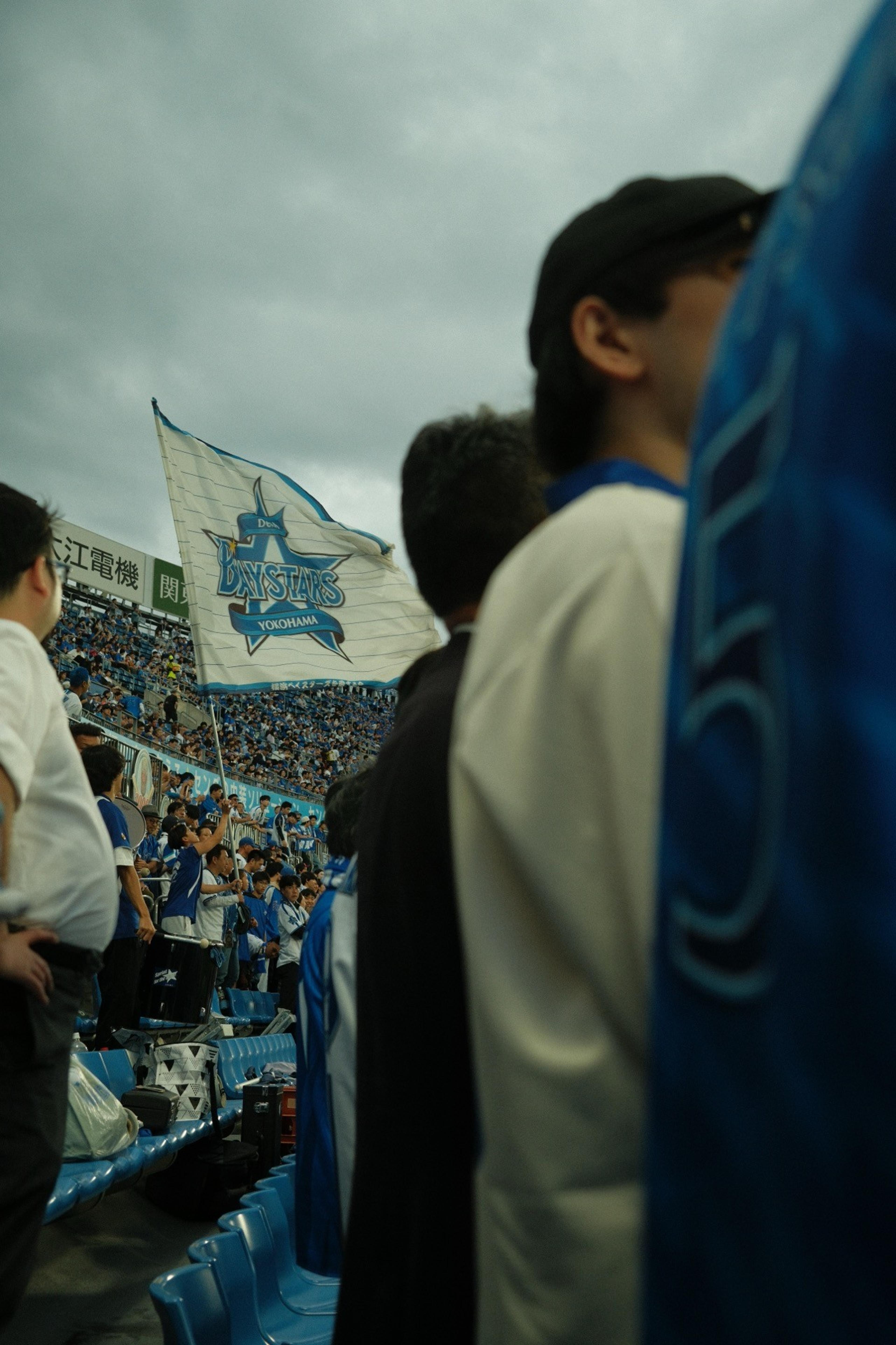 Fans in the stands under a cloudy sky as a Yokohama BayStars flag waves over the crowd