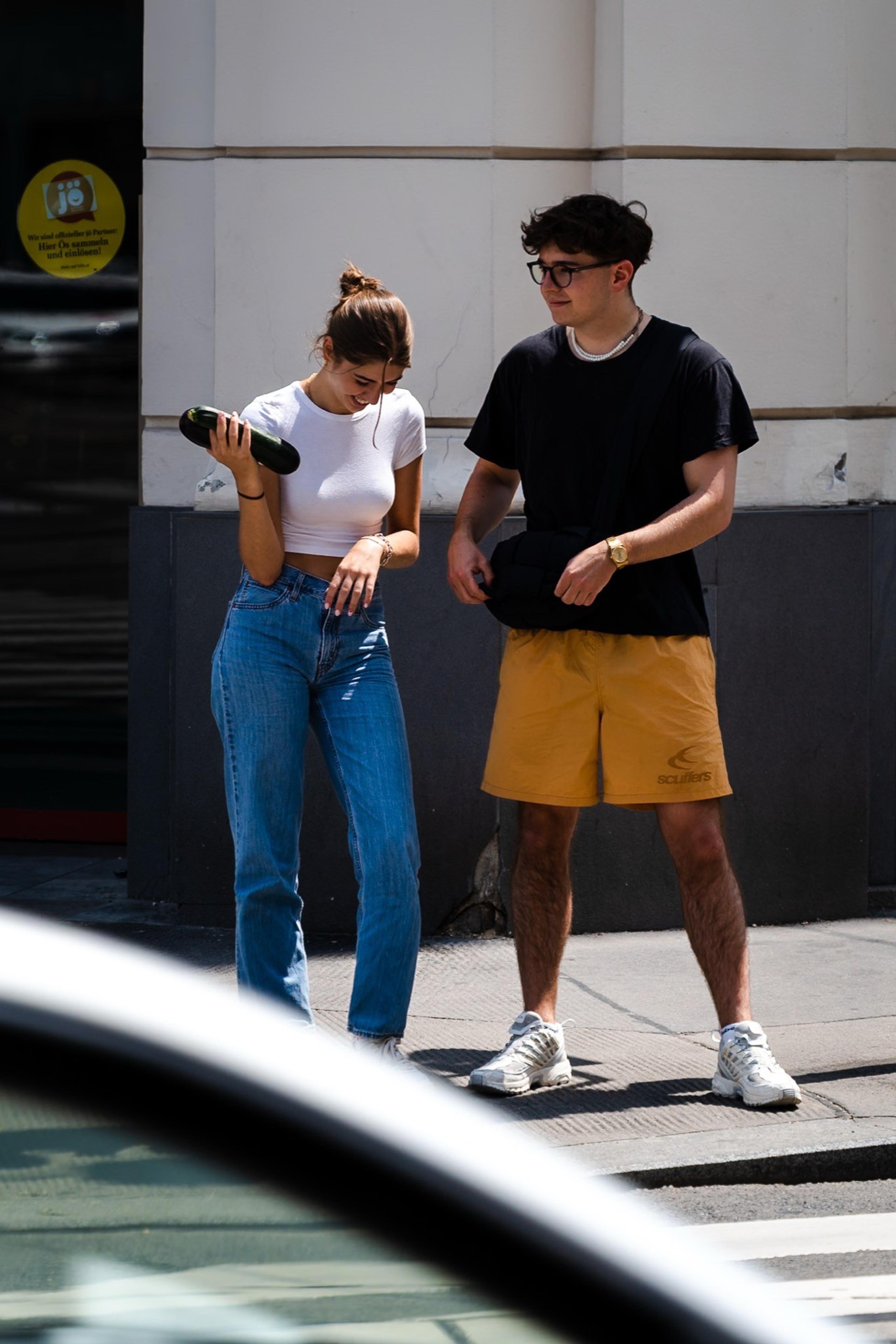Young couple laughing on a sunny street corner, she holds a sunglasses case, seen past a car