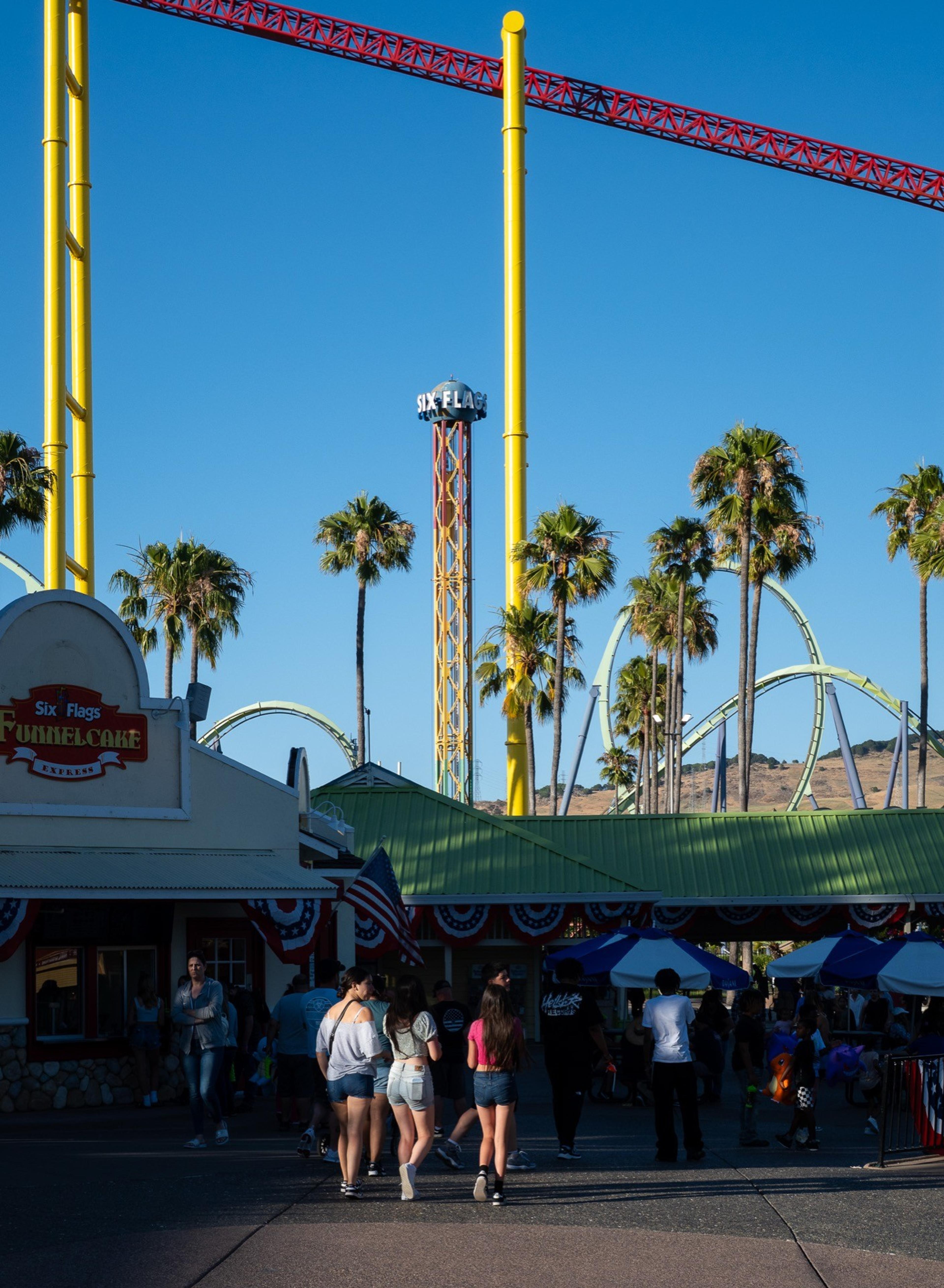 Visitors walking past a Six Flags funnel cake stand under roller coaster tracks and palm trees